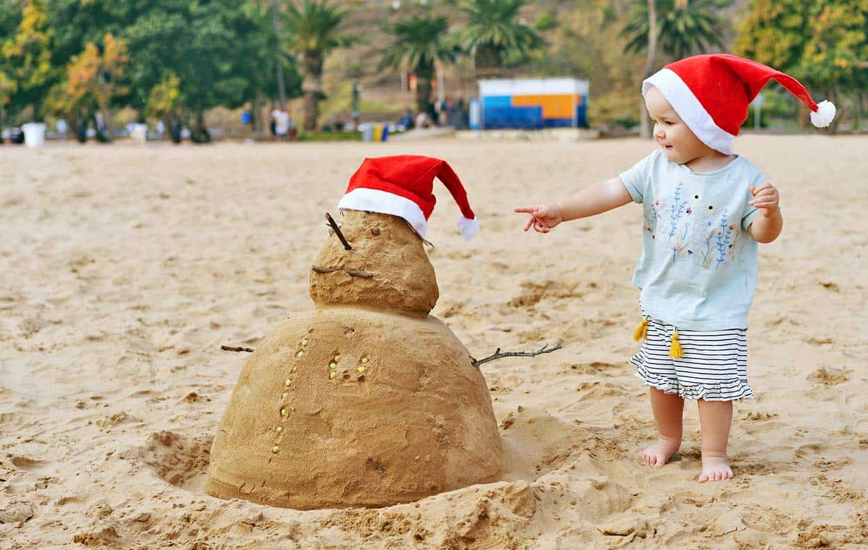 infant-boy-playing-in-sand-with-snowman-christmas-hat-at-beach-during-holiday-family-vacation-trip