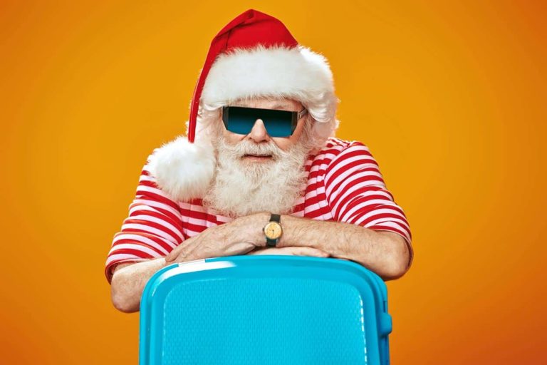 santa-with-shades-striped-shirt-waiting-to-go-on-holiday-vacation-with-blue-suitcase-sunset-orange-background