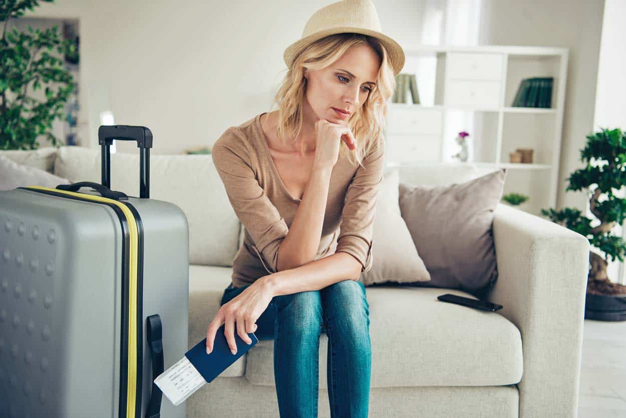 woman-in-hat-with-luggage-on-couch-looking-depressed-travel-plans-hindered-by-american-outbreak-social-distancing-order-plane-tickets-passport-in-hand
