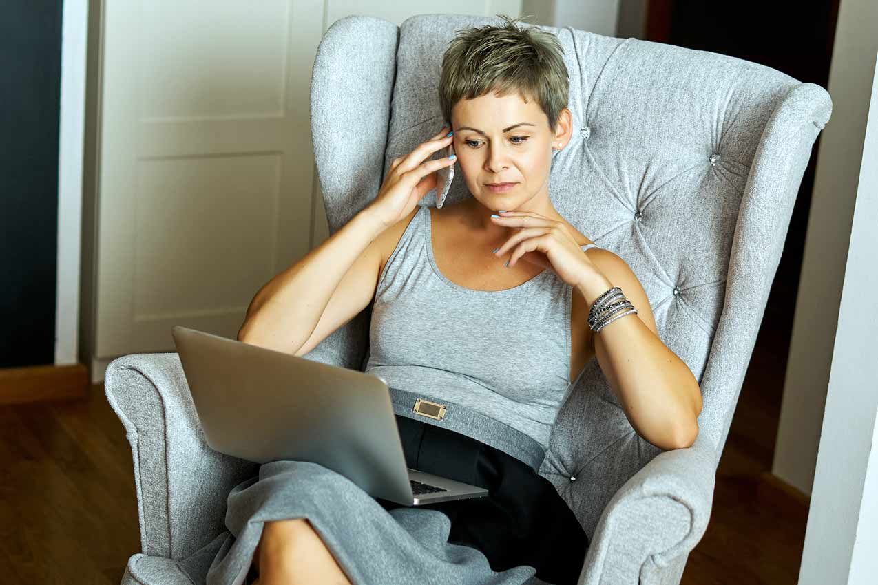 short-haird-woman-in-thinking-chair-on-phone-researching-internet-on-computer-at-home-during-pandemic