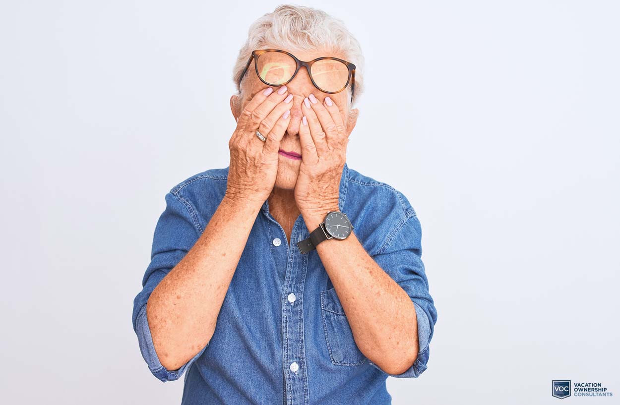 older-lady-rubbing-eyes-as-if-she-can't-believe-what-she's-seeing-and-hearing-from-timeshare-sales-presentation-white-background-denim-button-up-glasses-on-head