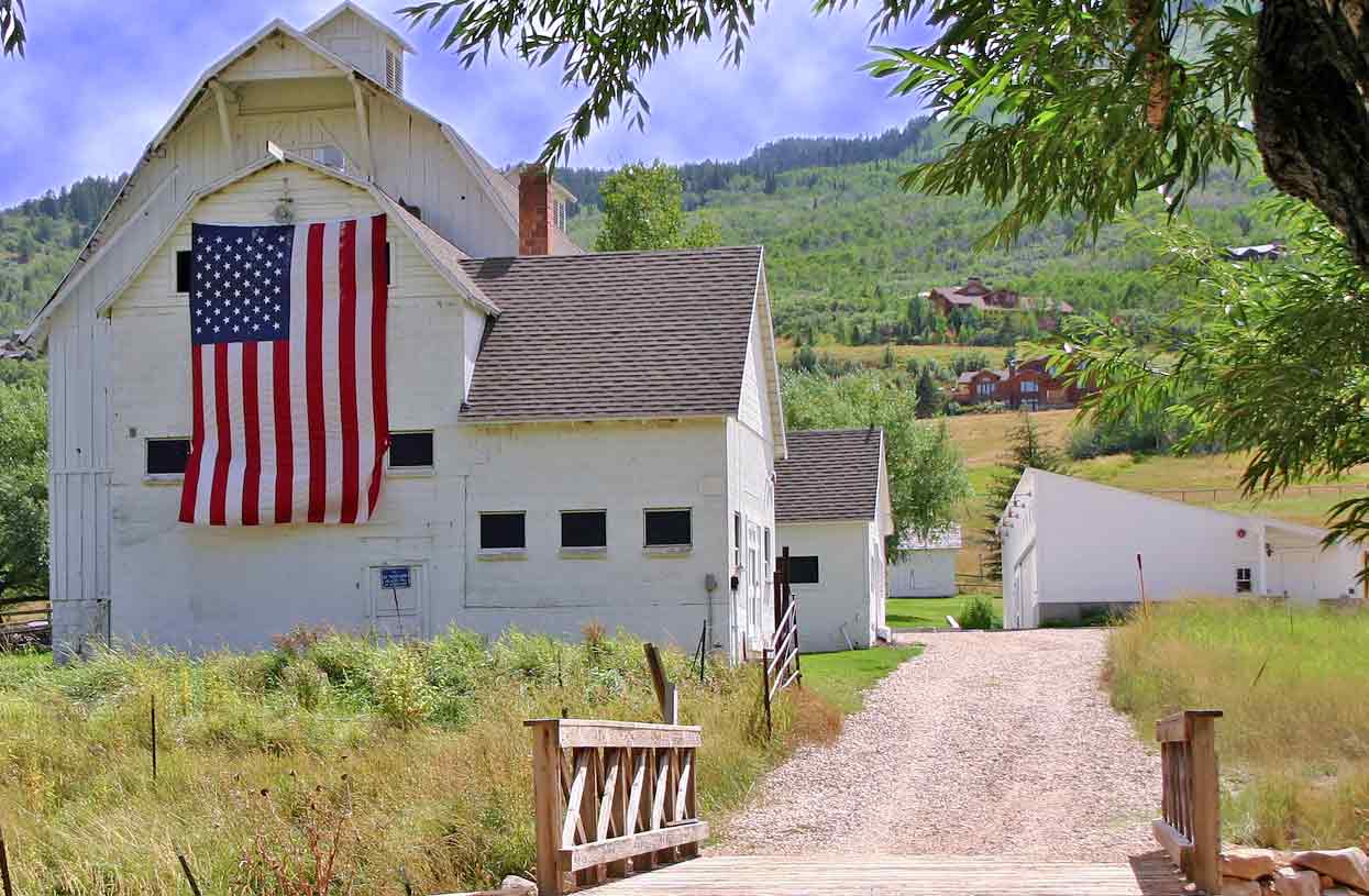 classic-western-frontier-in-utah-american-flag-on-white-washed-barn-hillside-drive-up-mountain-to-other-residencies-in-park-city-for-voc-blog