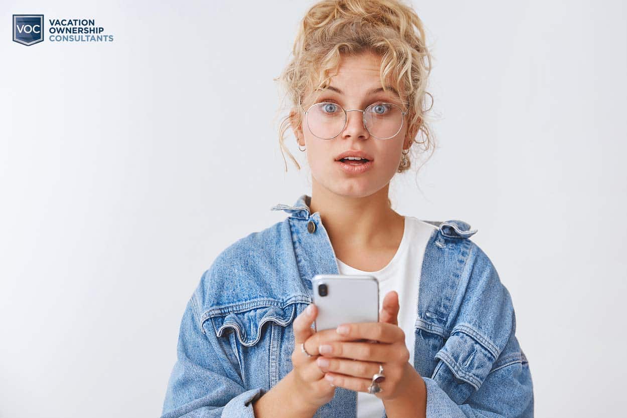 worried-looking-20-something-in-jean-jacket-white-backdrop-holding-cell-phone-wearing-glasses-for-timeshare-vs-homeownership-blog