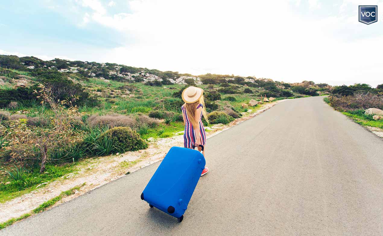 solo-female-tourist-walking-down-desert-road-greenery-with-bright-blue-luggage-with-wheels-wandering-for-vacation-during-covid-19-hoax