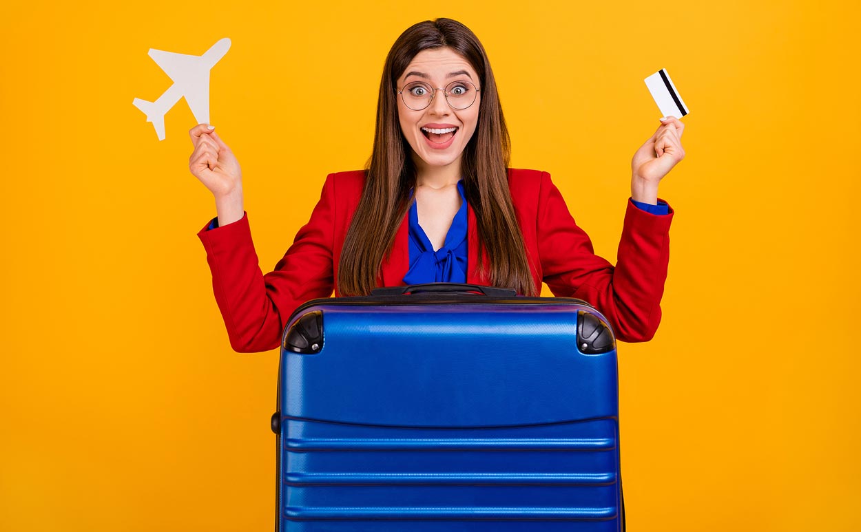 yellow-background-travel-gal-with-airplane-and-ticket-in-hand-behind-huge-blue-hard-cover-luggage-ready-to-go-excited