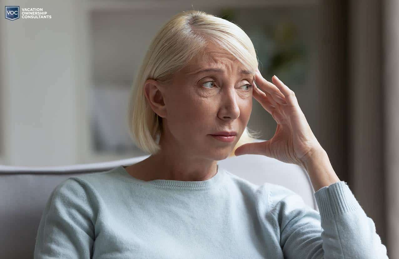 older blonde white woman pressing fingers against temple of forehead in worry as she contemplate canceling her timeshare or continuing to pay out of fear