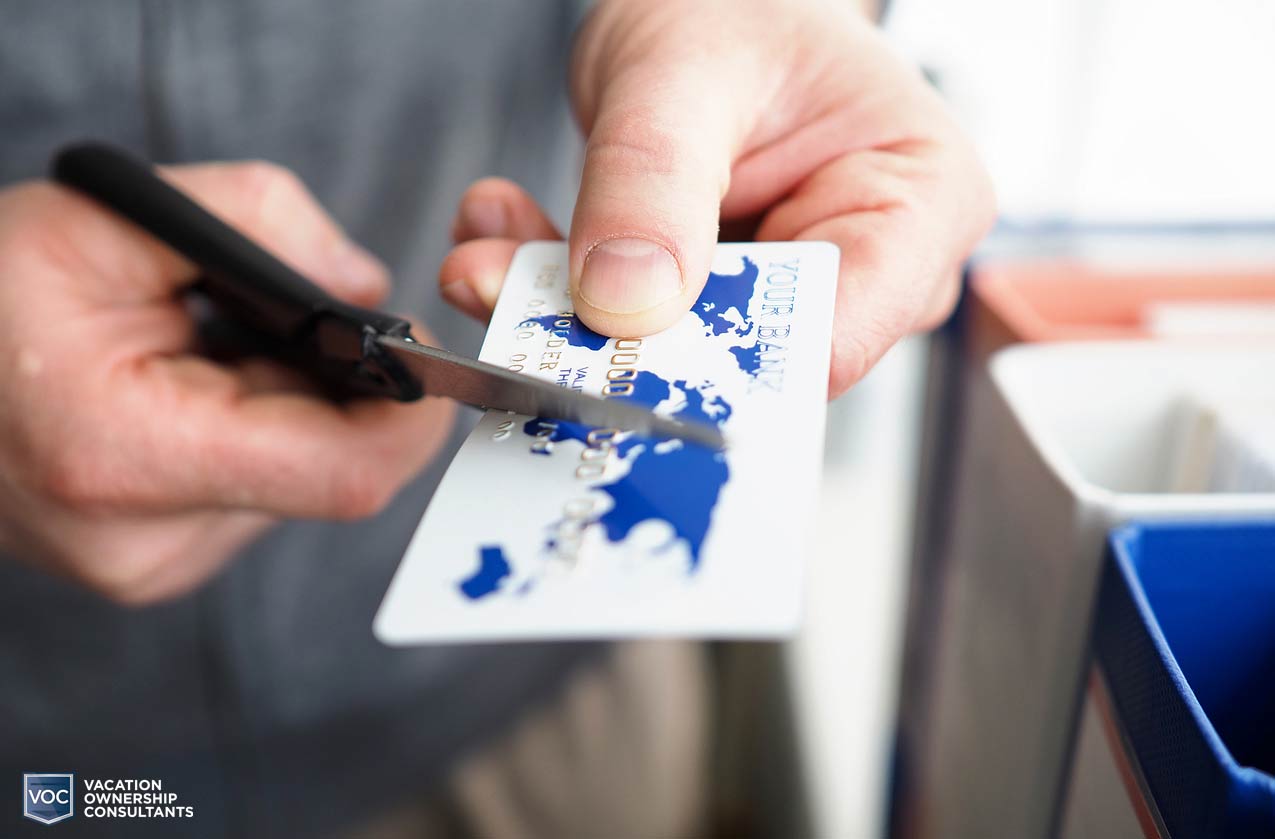 older hands of a man using black scissors to cut through a white credit card with blue land map on cover wearing grey sweatshirt and khakis on vacation