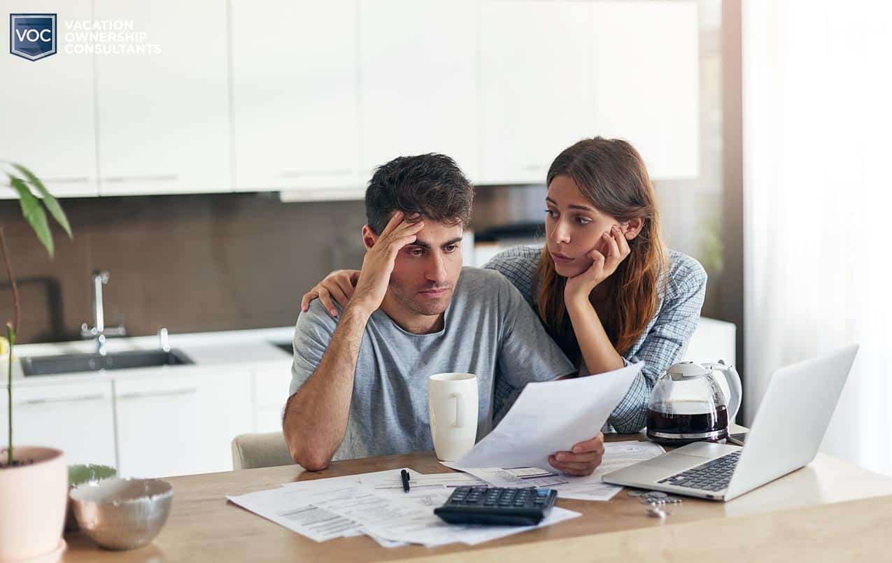 younger wife and husband couple in kitchen at table reading bills with large cup of coffee comforting him over timeshare bills by computer depressed