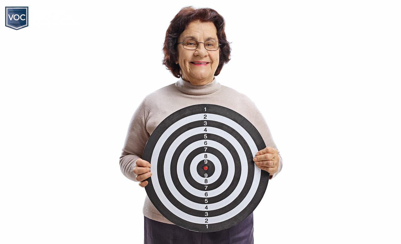 older woman dark hair brown outfit holding black and white target as if she is bullseye for sales practices white back drop voc